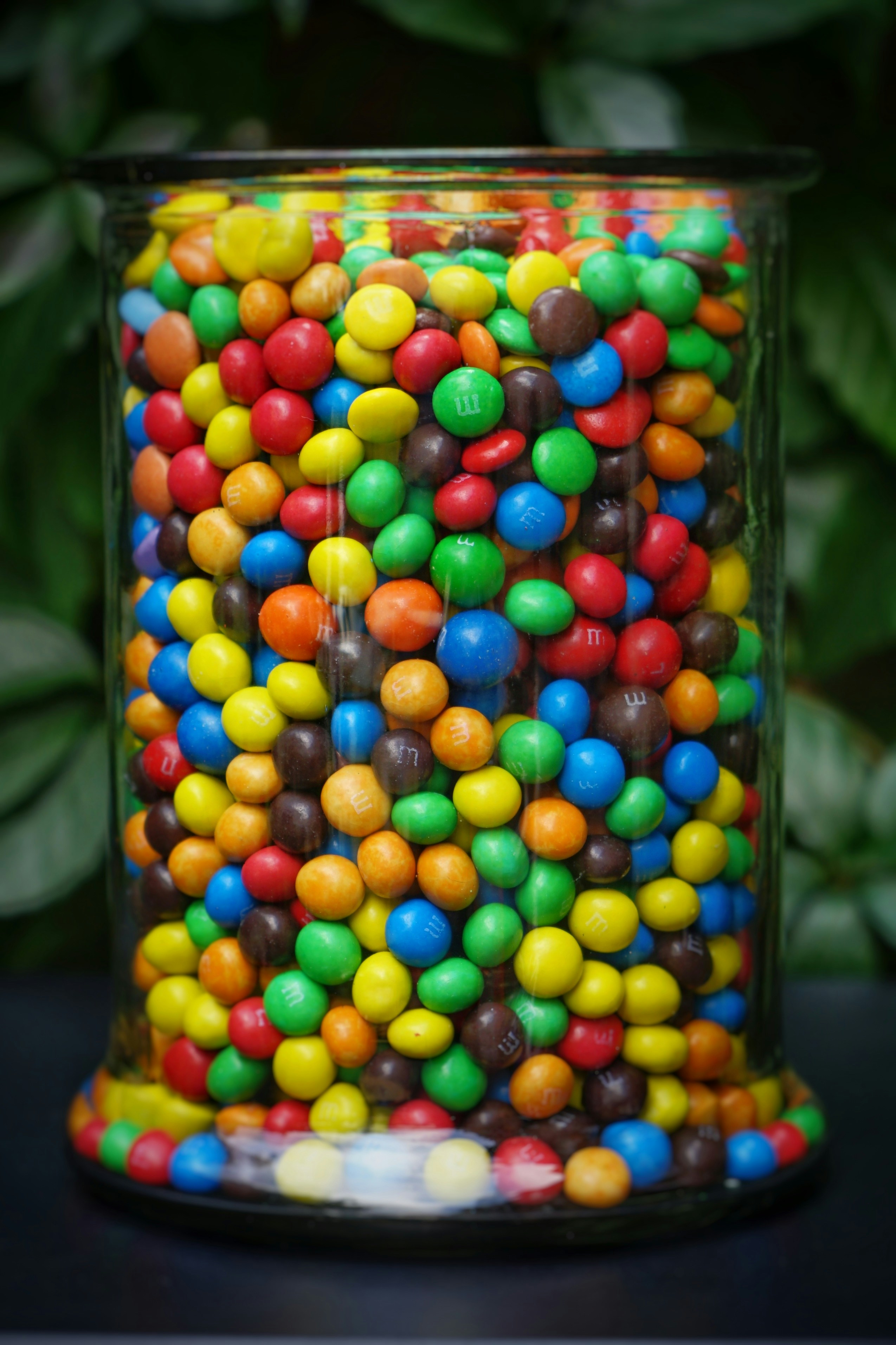 A tall glass jar packed with brightly colored candy, seen from the side so the mix of colors fills the jar.