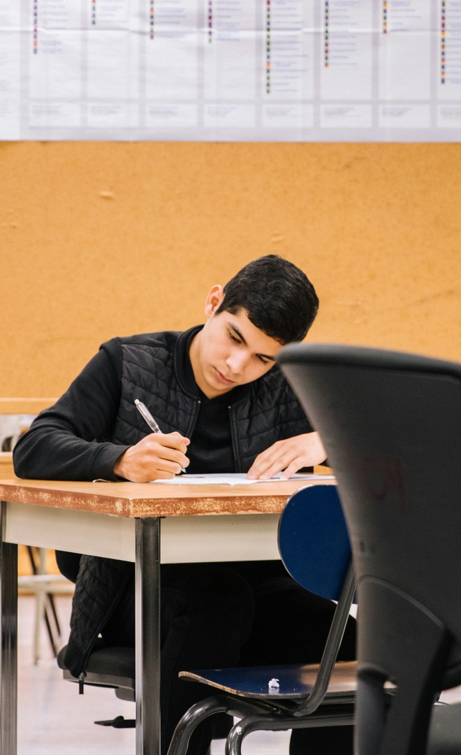 A student alone at a classroom desk, writing on a paper with a pen, in an otherwise empty room.