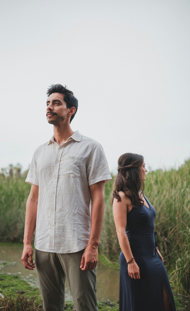 A man and a woman standing outdoors in tall grass under a pale overcast sky. The man is nearer the camera on the left and the woman a little behind him on the right; he is noticeably the taller of the two.