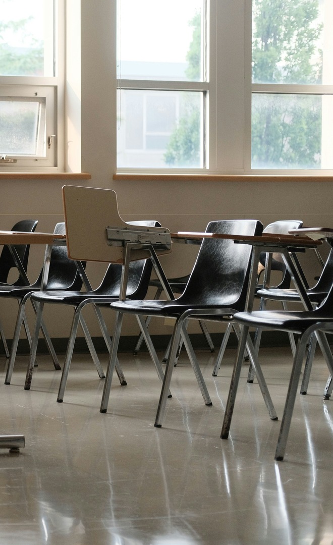 An empty classroom. Rows of black tablet-arm chairs stand unoccupied in front of tall daylit windows, with no students present.