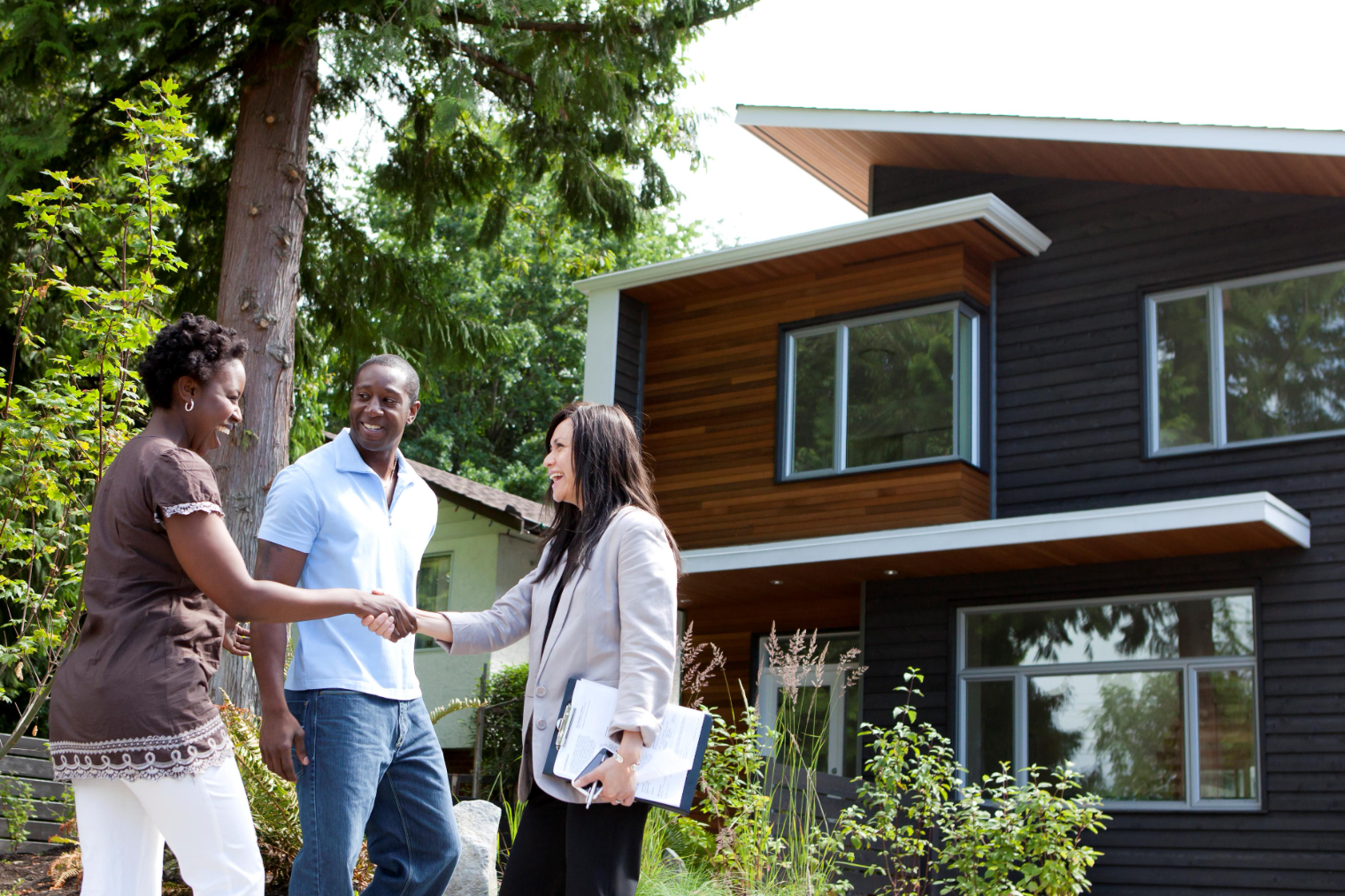 Two prospective buyers shake hands with an estate agent holding a clipboard, in front of a modern two-storey house with dark timber cladding.