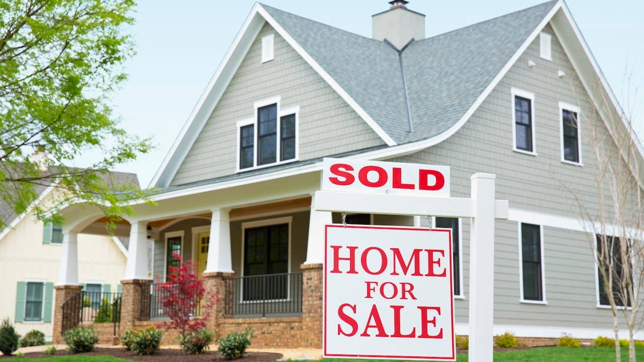 A gray clapboard house with a white porch behind a red-and-white HOME FOR SALE sign topped with a SOLD rider.
