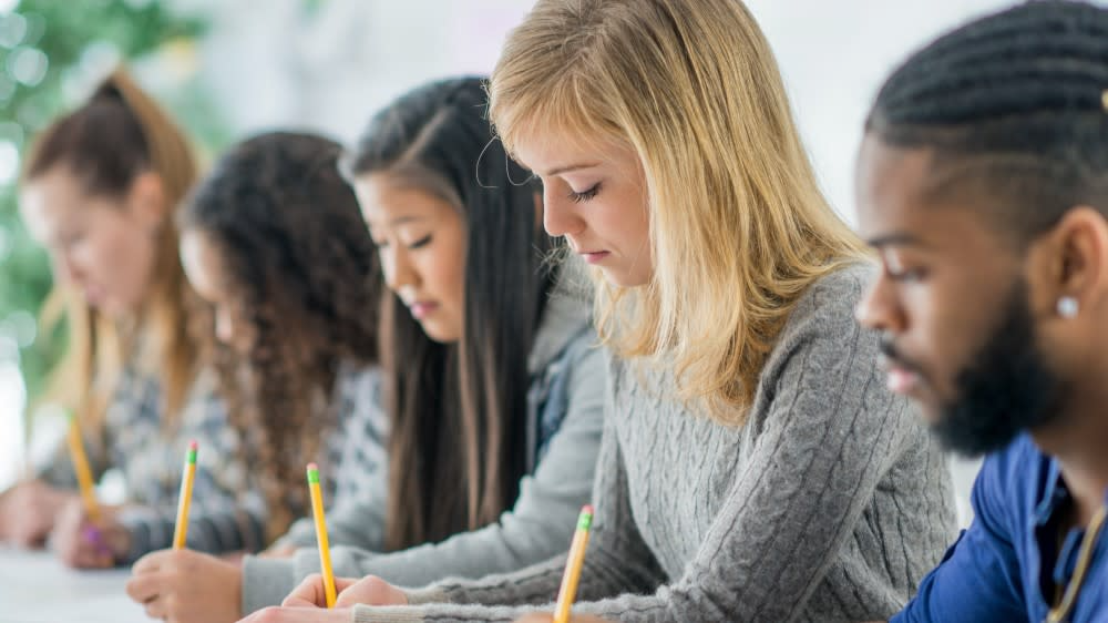 Five students seated in a row at a long table, heads down, writing with pencils.