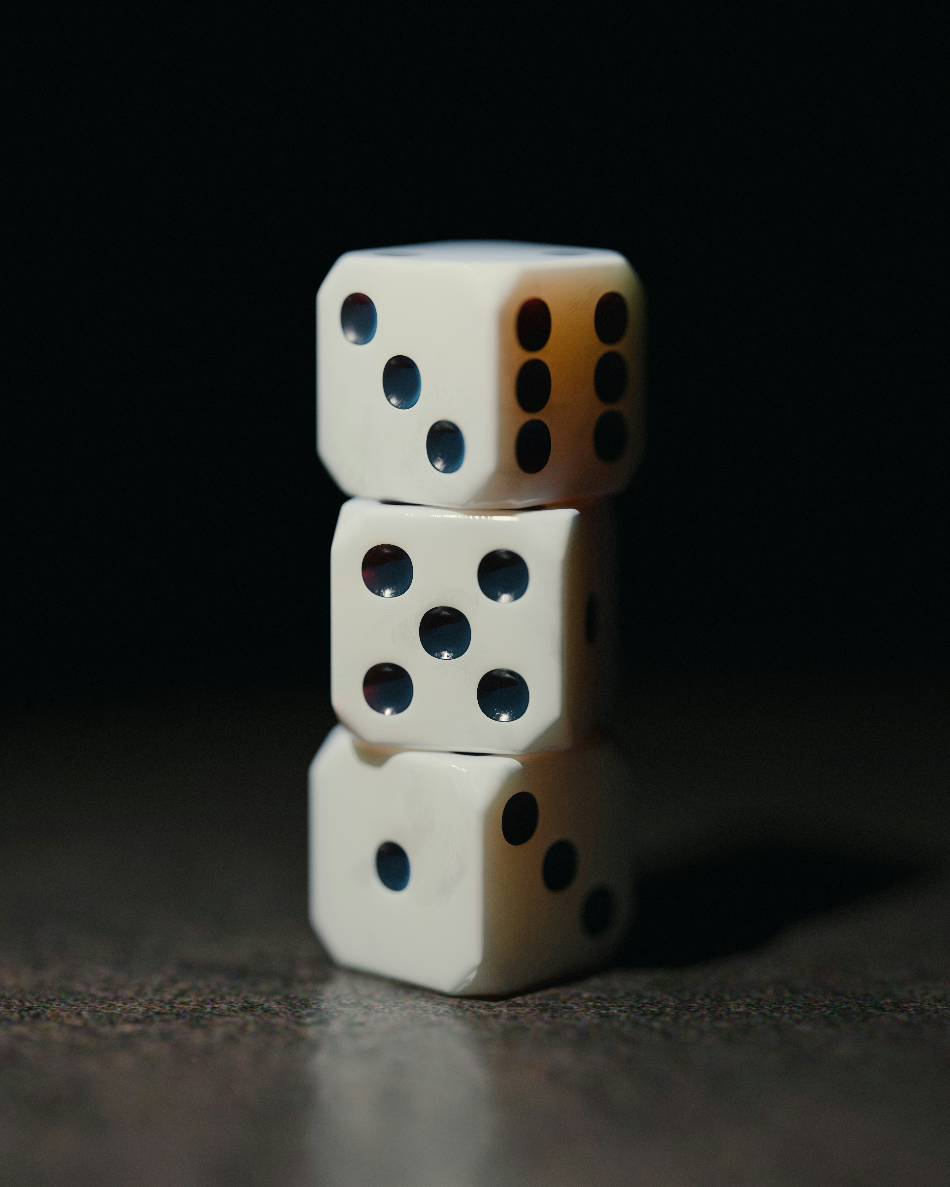 Three white dice stacked one on top of another on a dark surface, lit from the right against a black background. The top die shows three pips on the face toward the camera and six on its right side; the middle die shows five.