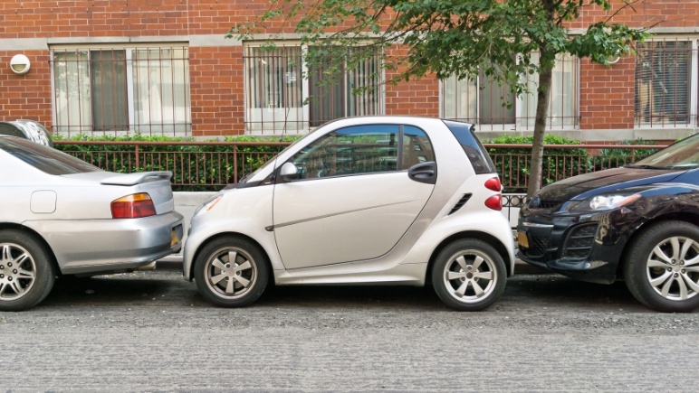 A small silver two-door city car parked at the curb between two larger cars, in front of a red-brick building.