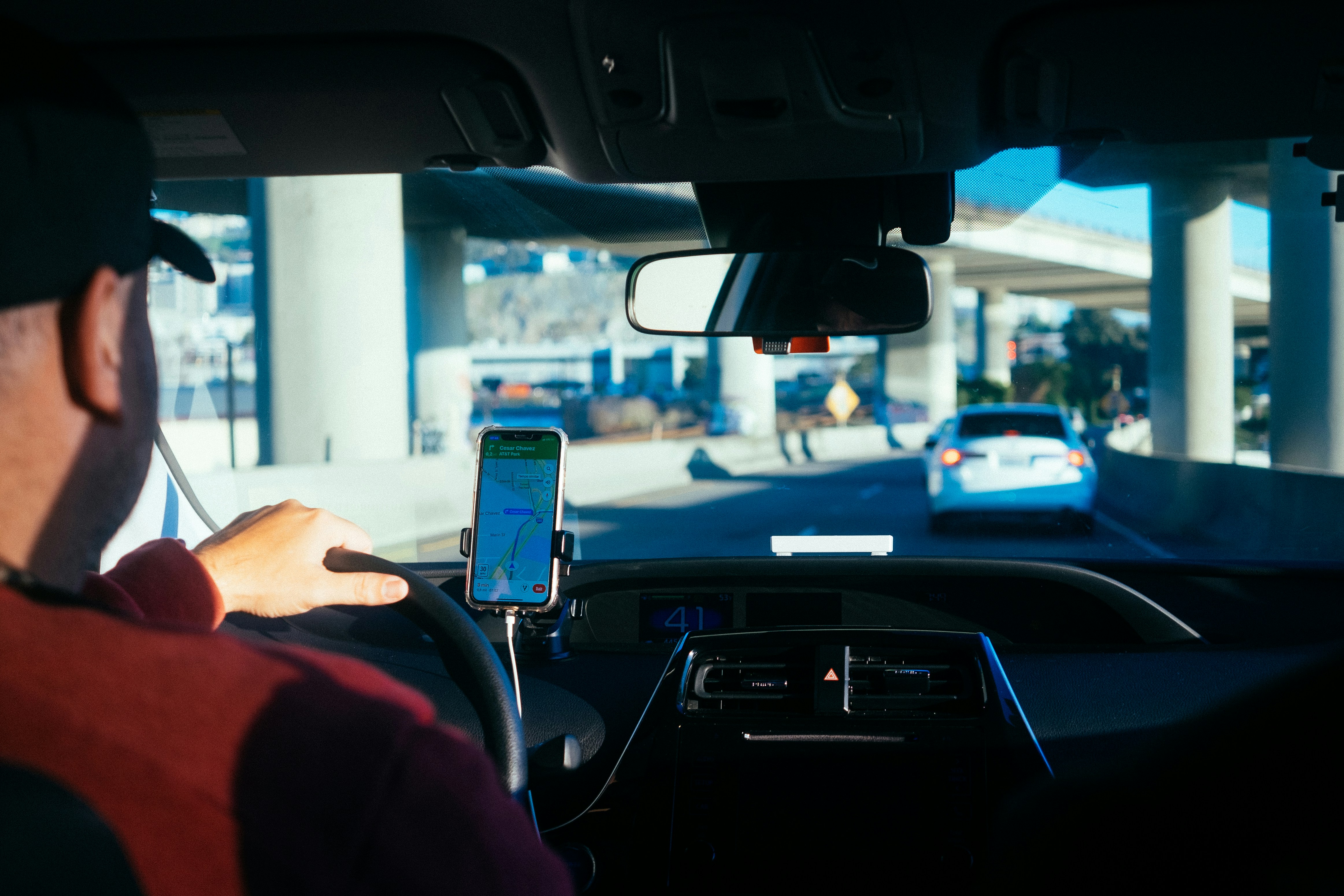 The view from a rideshare passenger seat: the driver in a cap and red hoodie with one hand on the wheel, a phone in a dashboard mount showing a navigation map, and freeway overpasses ahead through the windscreen.