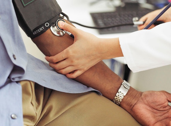 A clinician in a white coat presses a stethoscope to a patient’s inner elbow while a blood-pressure cuff is inflated on the upper arm.