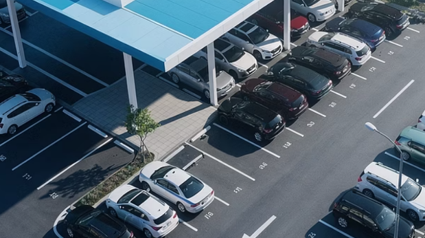 An aerial view of a parking lot. Rows of cars sit under blue-roofed canopies on one side and in open bays on the other, with directional arrows painted on the asphalt.