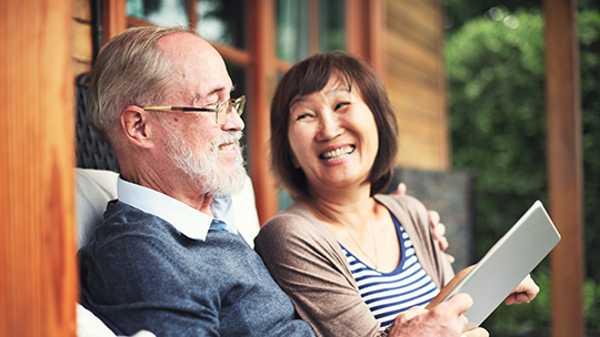 An older couple sit outdoors together, both smiling; the woman holds a tablet.