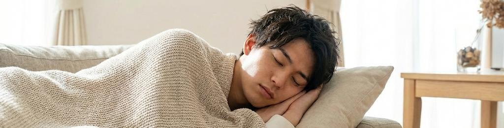 A young man asleep on a sofa under a knitted blanket.