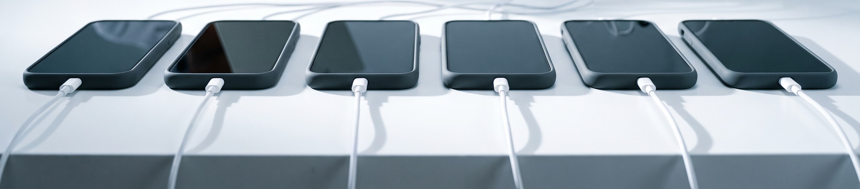 Six identical smartphones lie face up in a row on a test bench, each plugged into its own charging cable.