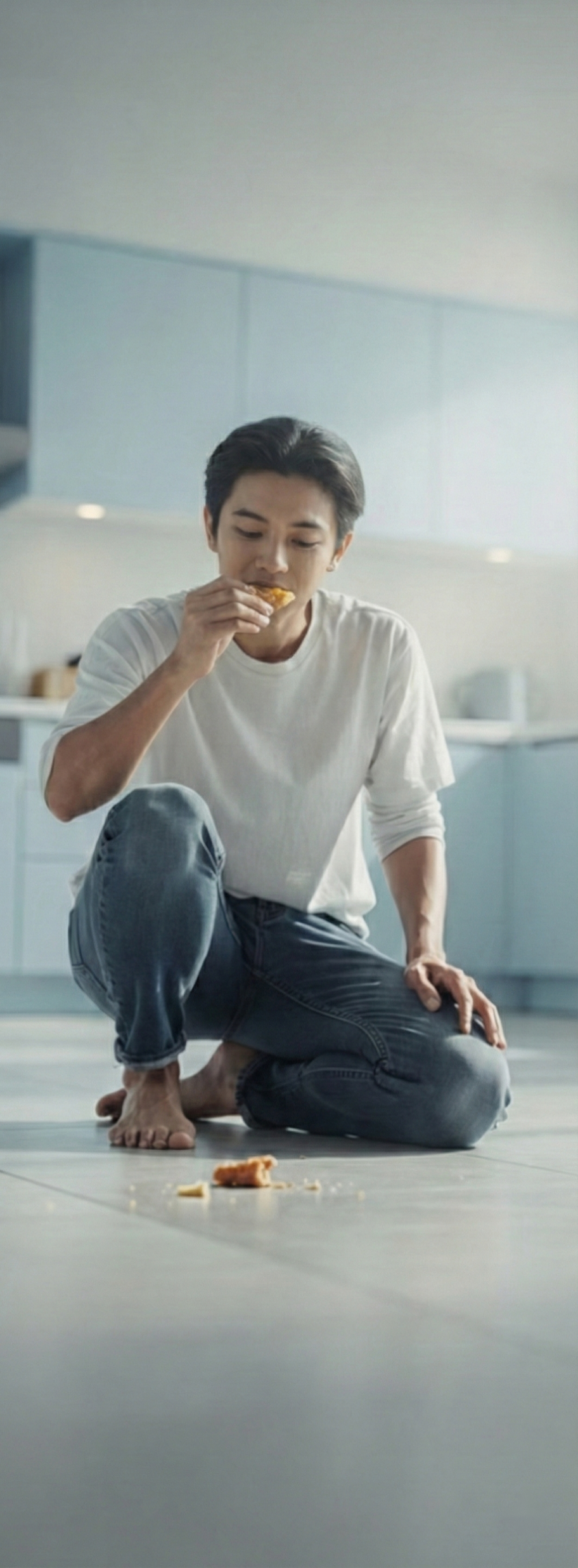 A young man kneels on one knee, barefoot, on a kitchen floor, eating a piece of food he has picked up off it, with crumbs scattered on the tiles in front of him.